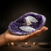 Hand holding a large purple amethyst crystal with visible white crystals inside against a neutral background