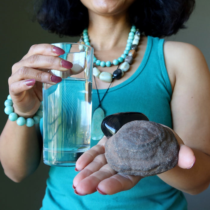 Person holding a glass of water and moqui marble and obsidian stone, wearing amazonite jewelry.