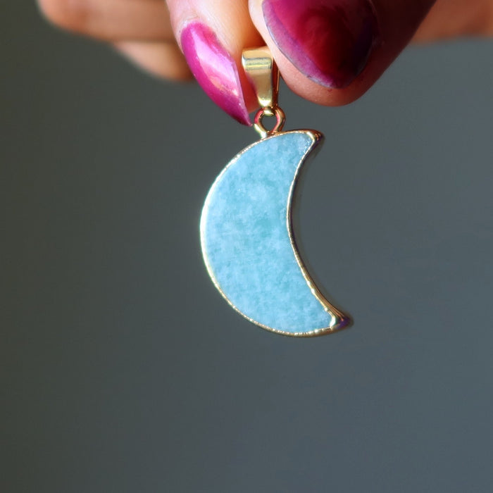 Blue Amazonite crescent moon pendant held by a hand with pink nail polish against a gray background