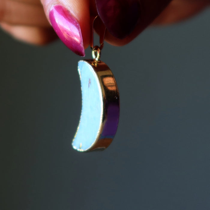Crescent moon-shaped Amazonite pendant with gold rim held by a hand with pink nail polish against a dark background