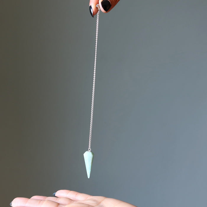 Light blue amazonite pendulum on a chain held by a hand against a gray background