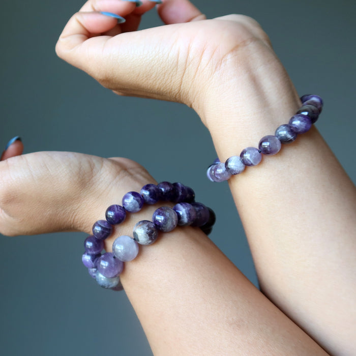 Two hands wearing chevron amethyst purple beaded bracelets against a gray background