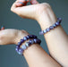 Two hands wearing chevron amethyst purple beaded bracelets against a gray background