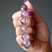 Hand holding an amethyst necklace with purple and clear beads against a neutral background