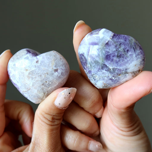 Two heart-shaped purple and white amethyst stones held between fingers against a neutral background