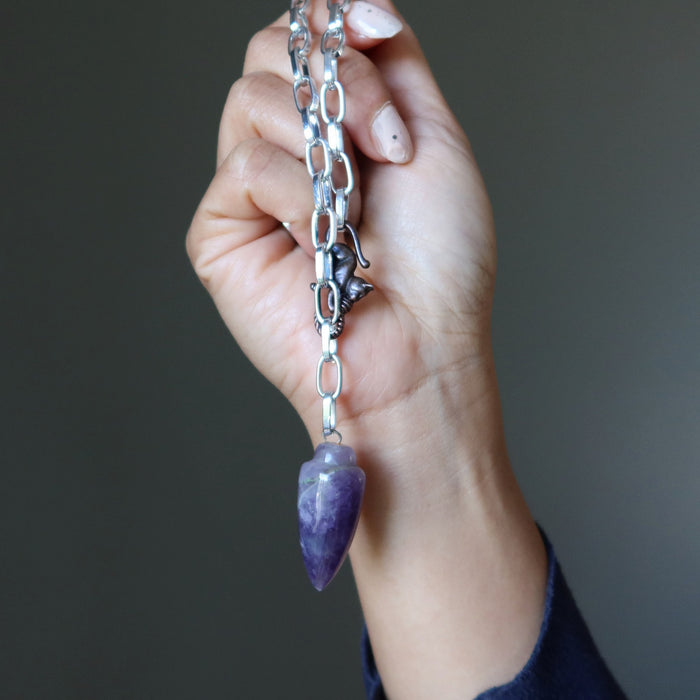Hand holding a silver chain with a purple amethyst crystal pendant against a dark background