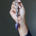 Hand holding a silver chain with a purple amethyst crystal pendant against a dark background