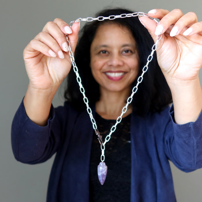 Person holding a silver chain necklace with a purple amethyst pendant against a gray background