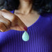 Person holding a light green Aventurine teardrop-shaped pendant against a blurred purple background