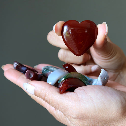 Hand holding a heart-shaped red stone with other stones on a gray background