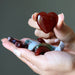 Hand holding a heart-shaped red stone with other stones on a gray background