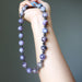 Hand holding a charoite beaded necklace with purple and black beads against a gray background