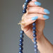 Hand holding a blue dumortierite beaded necklace with a gold clasp against a gray background