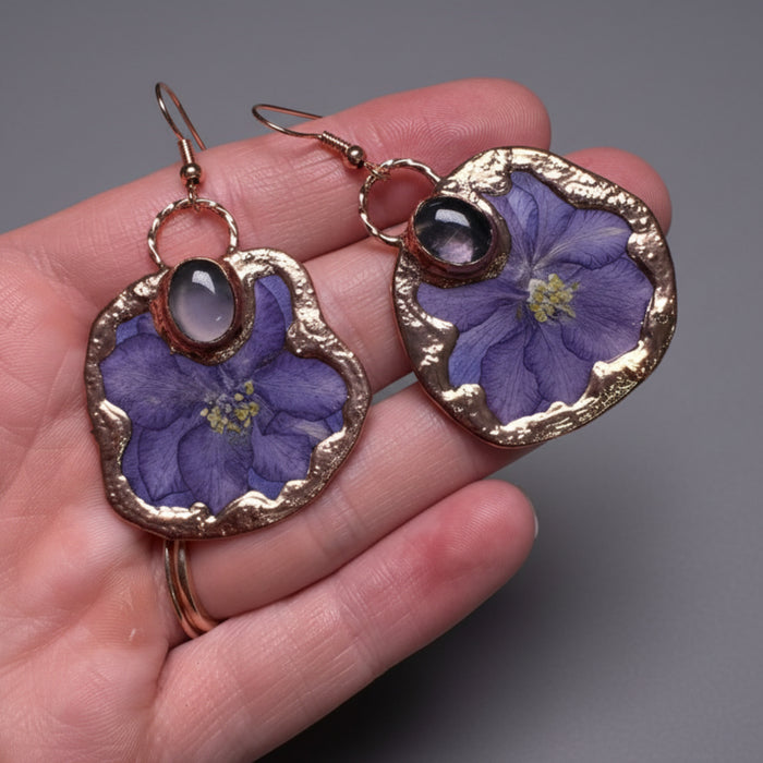 Earrings with purple floral design and gold accents held in a hand against a gray background
