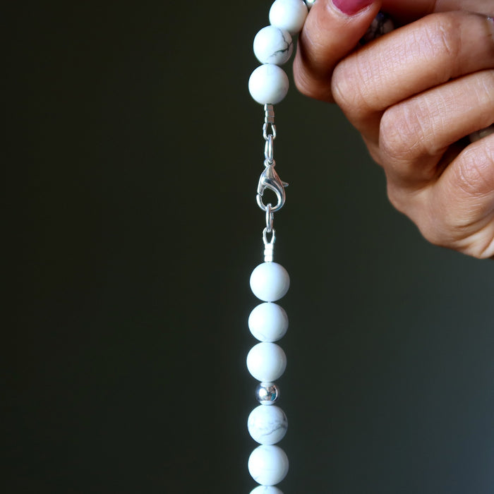 Hand holding a Howlite necklace with white beads against a dark background