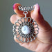Silver floral Howlite pendant held by a hand with pink nail polish against a neutral background