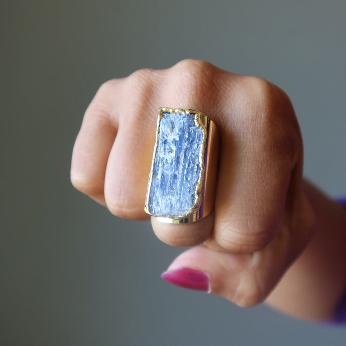 Hand wearing a ring with a blue Kyanite stone and gold band on a neutral background
