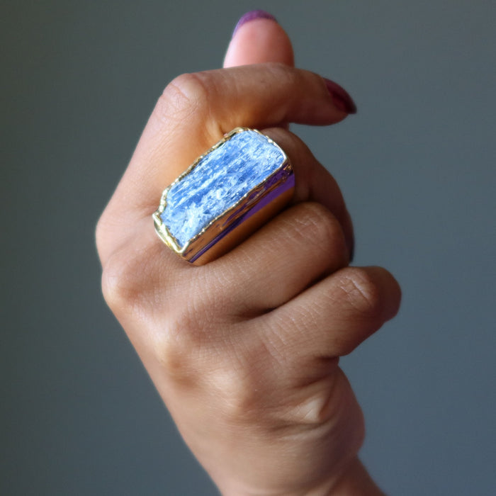 Hand wearing a ring with a blue Kyanite crystal design on a gray background