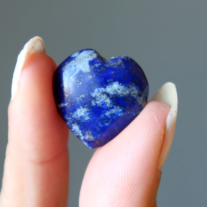 Heart-shaped blue lapis stone held between fingers against a gray background