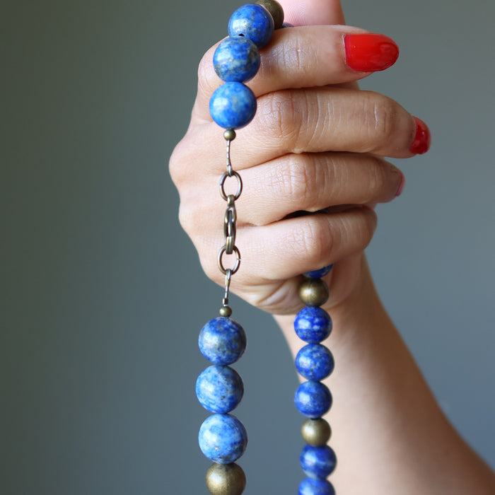 Hand holding a beaded Lapis Lazuli necklace with blue and gold beads against a gray background