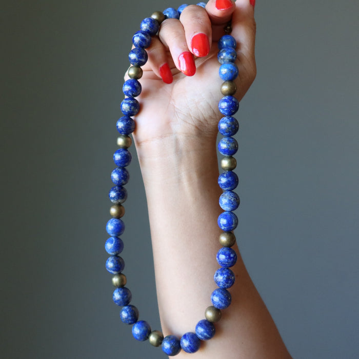 Hand holding a blue beaded Lapis Lazuli necklace against a gray background