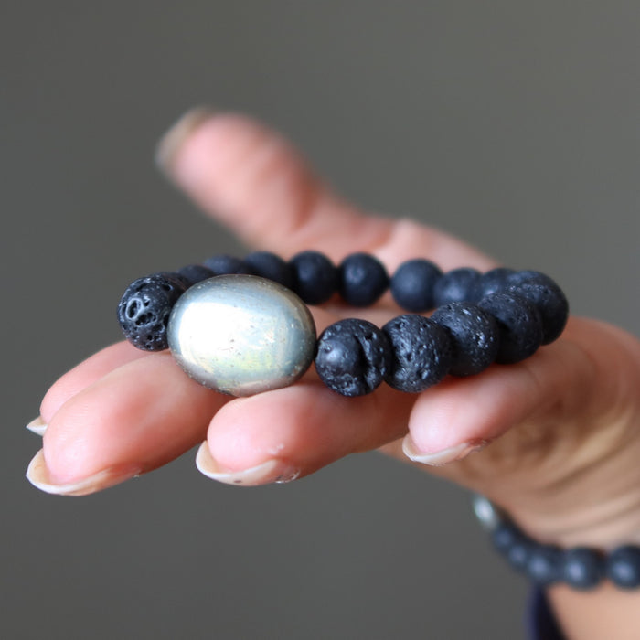 Hand holding a bracelet with lava beads and a large pyrite bead against a neutral background