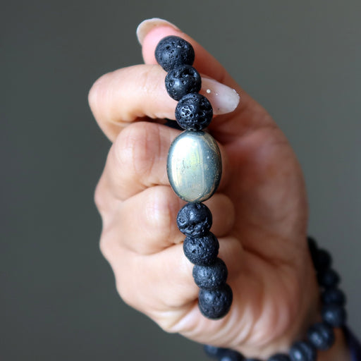 Hand holding a bracelet with black lava beads and a central pyrite bead against a neutral background
