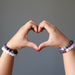 Hands making a heart shape with purple amethyst and pink rose quartz beaded bracelets on a gray background