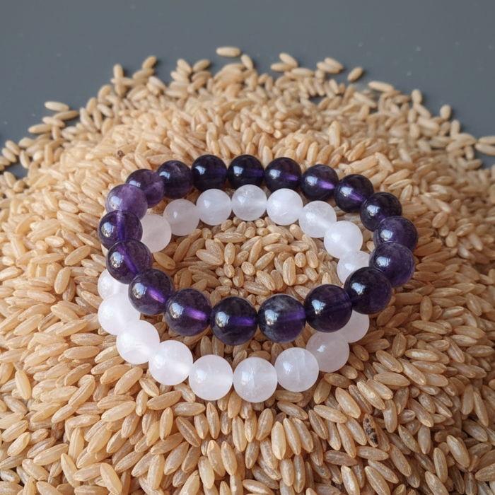 Two beaded bracelets, one amethyst purple and one rose quartz pink, on a bed of rice.