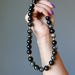 Hand holding a black beaded Gold Sheen Obsidian necklace against a gray background
