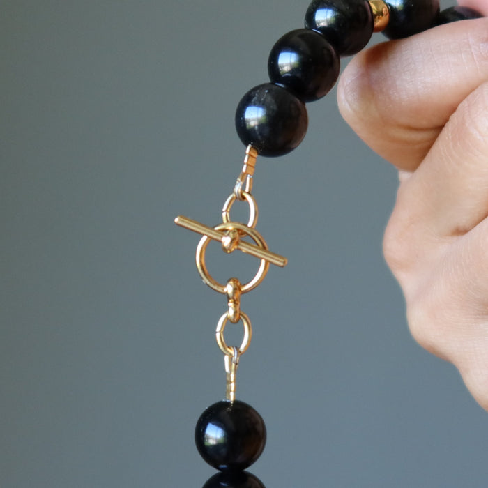 Black beaded Gold Sheen Obsidian necklace with a gold clasp held by a hand against a gray background