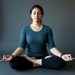 Woman in a teal shirt and black pants sitting in a meditative pose on a gray background