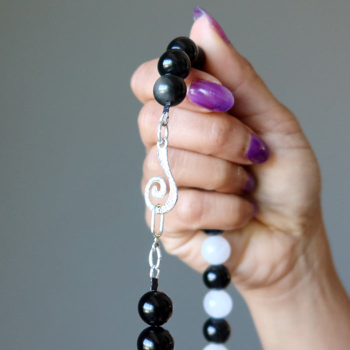 Hand holding a black and white beaded Rainbow Obsidian Snow Quartz necklace with a silver clasp against a gray background