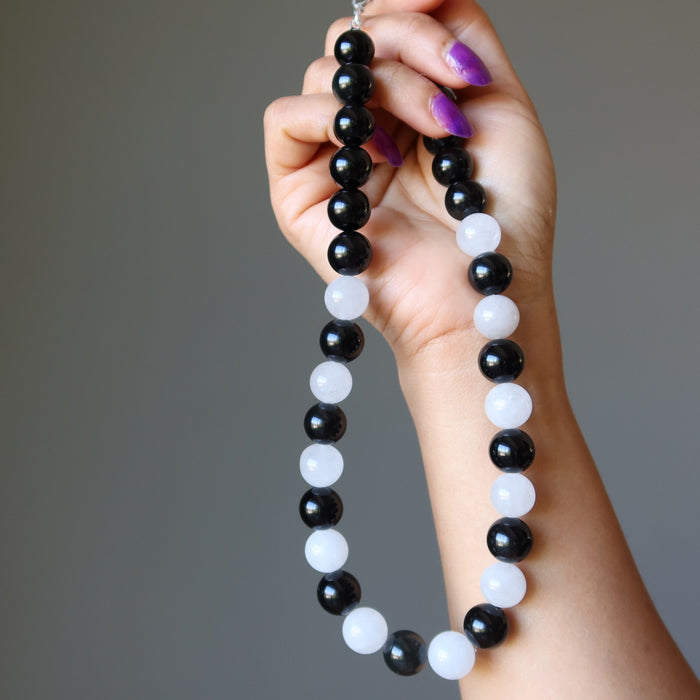 Hand holding a black and white beaded Rainbow Obsidian Snow Quartz necklace against a gray background