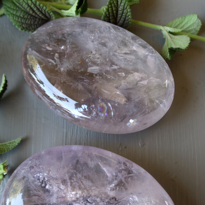 Two Amethyst crystal stones on a light wooden surface with green leaves in the background