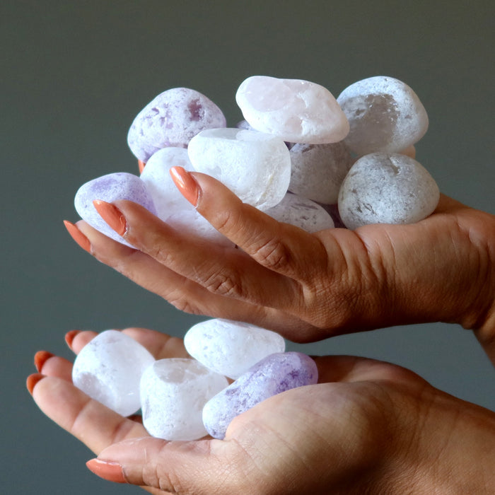 Hands holding a pile of small, round, quartz stones with purple accents against a neutral background