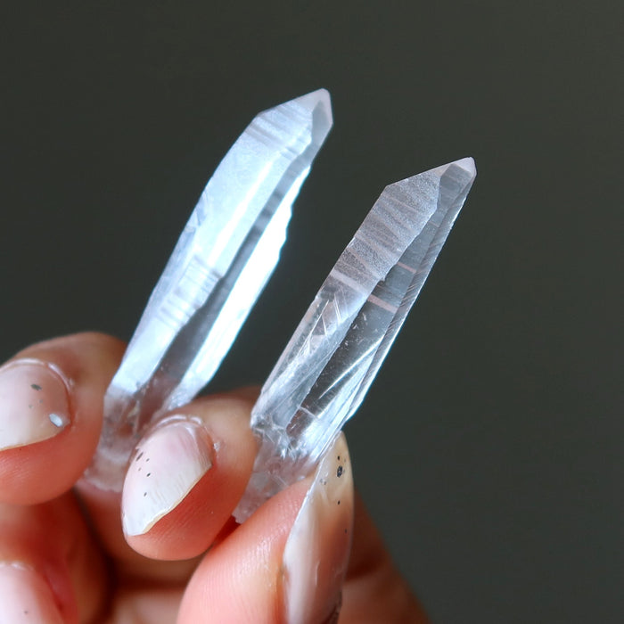 Two clear lemurian quartz point crystal held between fingers against a dark background