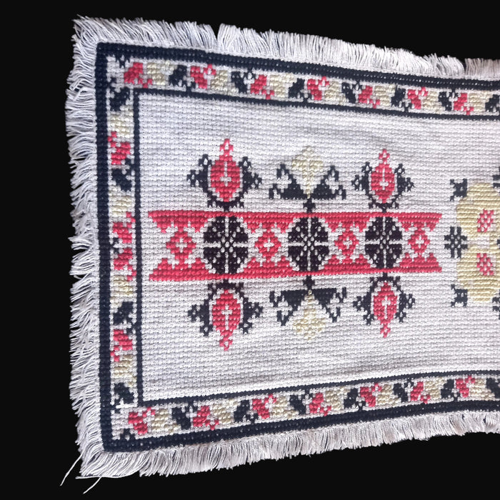 Close up of a Hungarian table runner in beregi style showing red diamonds and black geometric stars, with a fringed border.