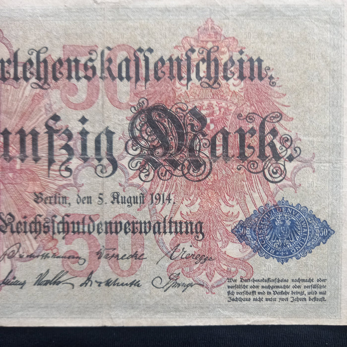 A vintage German 50 Reichsbanknote from August 5, 1914, displayed flat against a dark navy background. The note features a cream-colored paper base with red and blue printed details, black gothic text, and ornate red floral patterns.