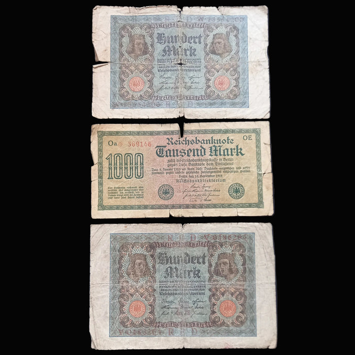 Three vintage German banknotes laid flat on a dark fabric background, arranged vertically. The top note is a Burdert Mark, the middle is a 1000 Mark note, and the bottom is a larger Mark note.