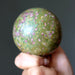 Hand holding a green and pink speckled ruby fuchsite sphere against a dark background