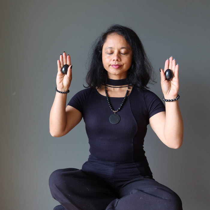 Woman in a meditative pose holding black Shungite stones against a gray background