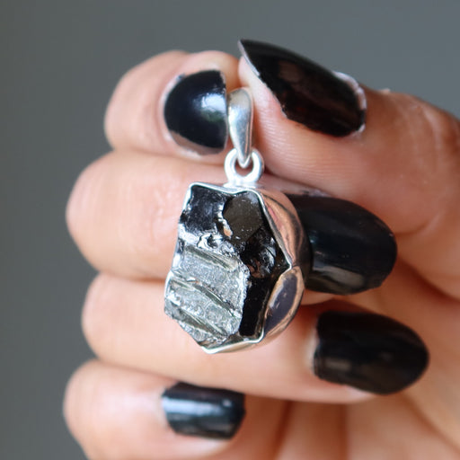 Hand holding a silver pendant with a rough-cut  Elite Shungite gemstone against a neutral background