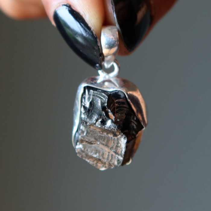 Close-up of a silver pendant with a  Elite Shungite stone held by a hand with black nail polish.