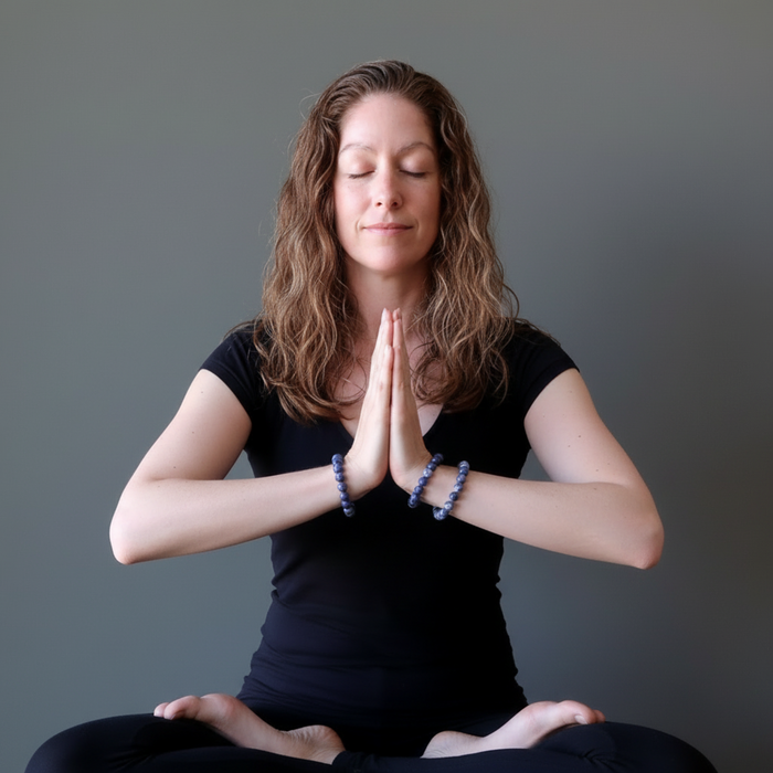 Woman in a black outfit sitting in a meditative pose with hands pressed together against a gray background