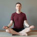 Man in maroon t-shirt and black shorts sitting in a yoga pose on a mat with a neutral background