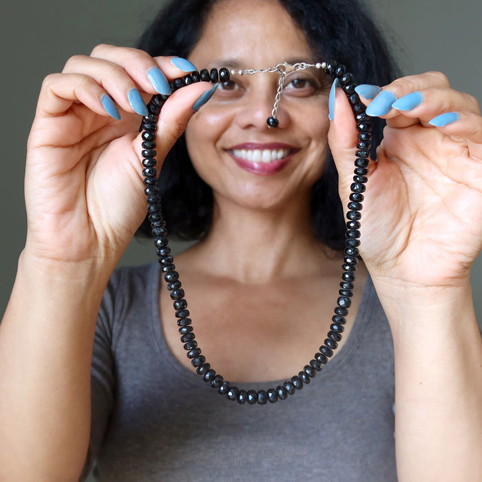 Woman holding a black Tourmaline beaded necklace in front of her face against a neutral background