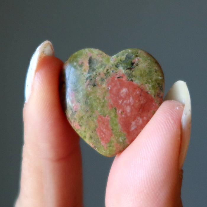 Heart-shaped multicolored unakite stone held between fingers against a neutral background