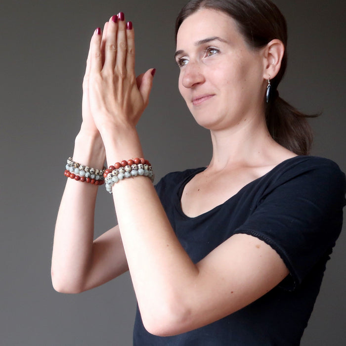 lucia of satin crystals with hands in prayer wearing set of 3 sesame, red and dalmatian jasper stretch bracelets on each hand.